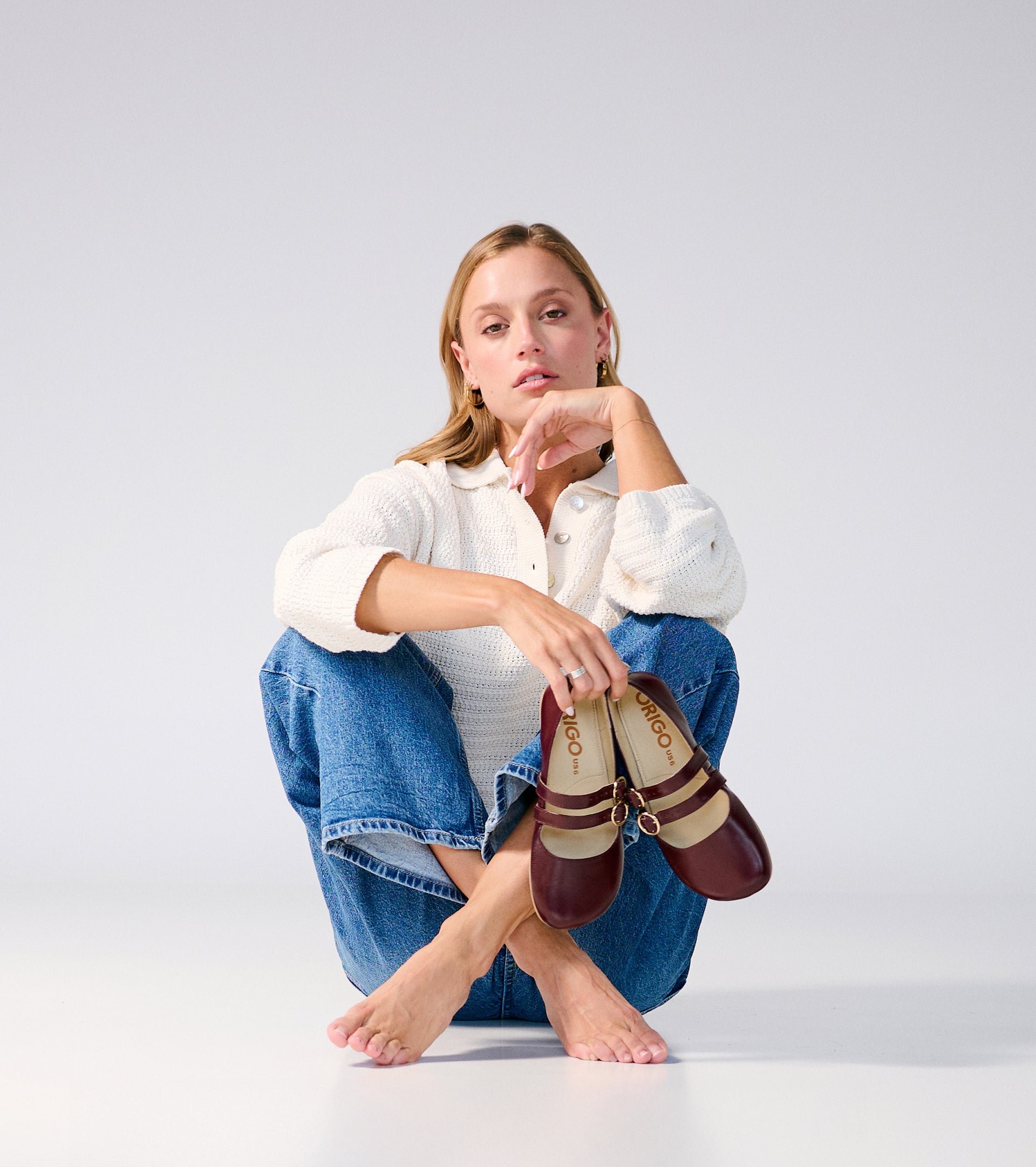 Woman sitting on the floor holding a pair of patent burgundy barefoot flats with a white background