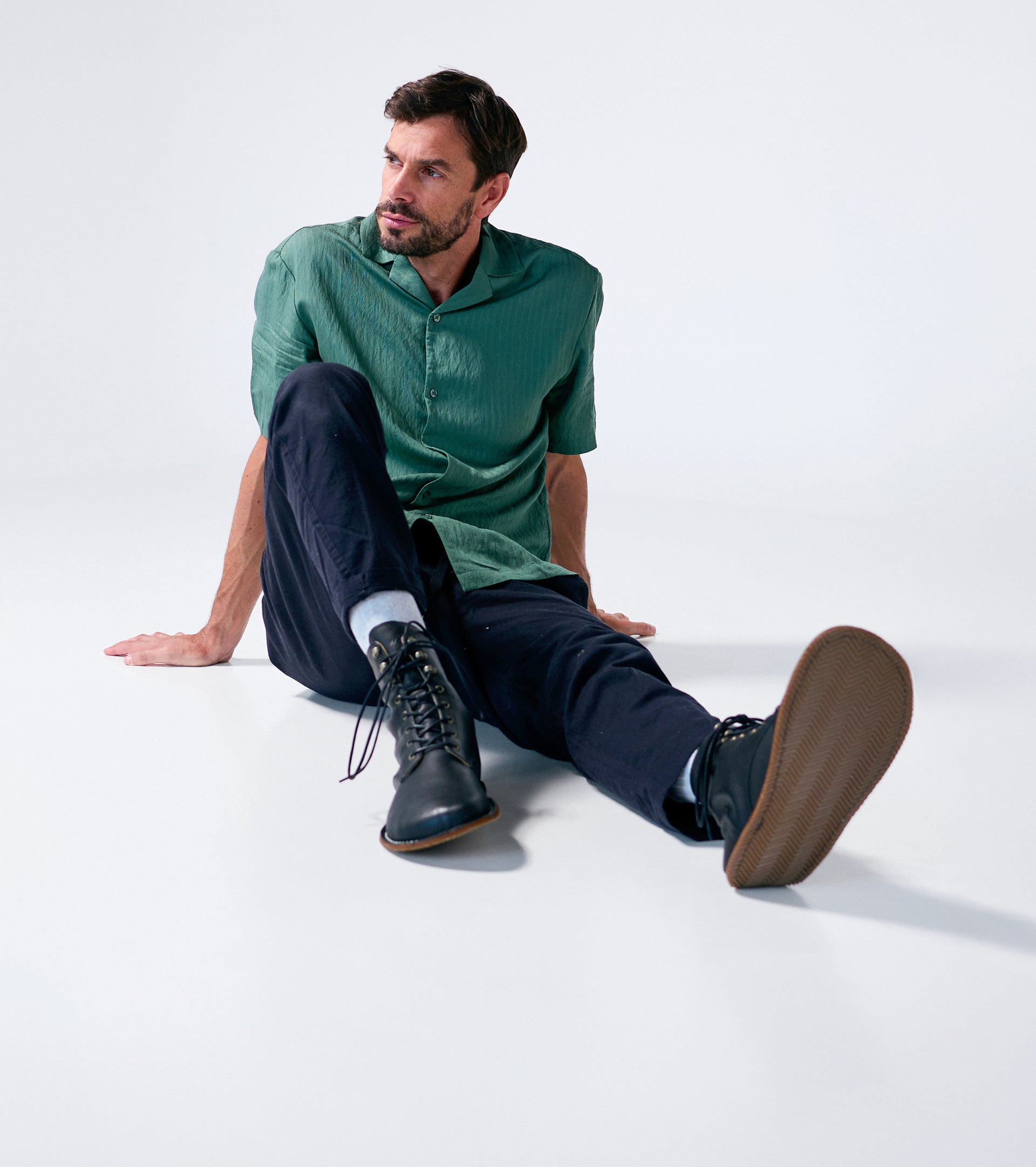 Man sitting on a white floor wearing a green shirt and dark pants and black barefoot boots