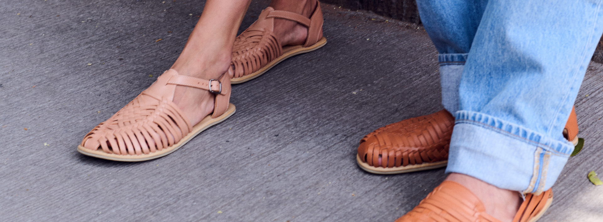 Two pairs of feet wearing brown sandals on a gray pavement.